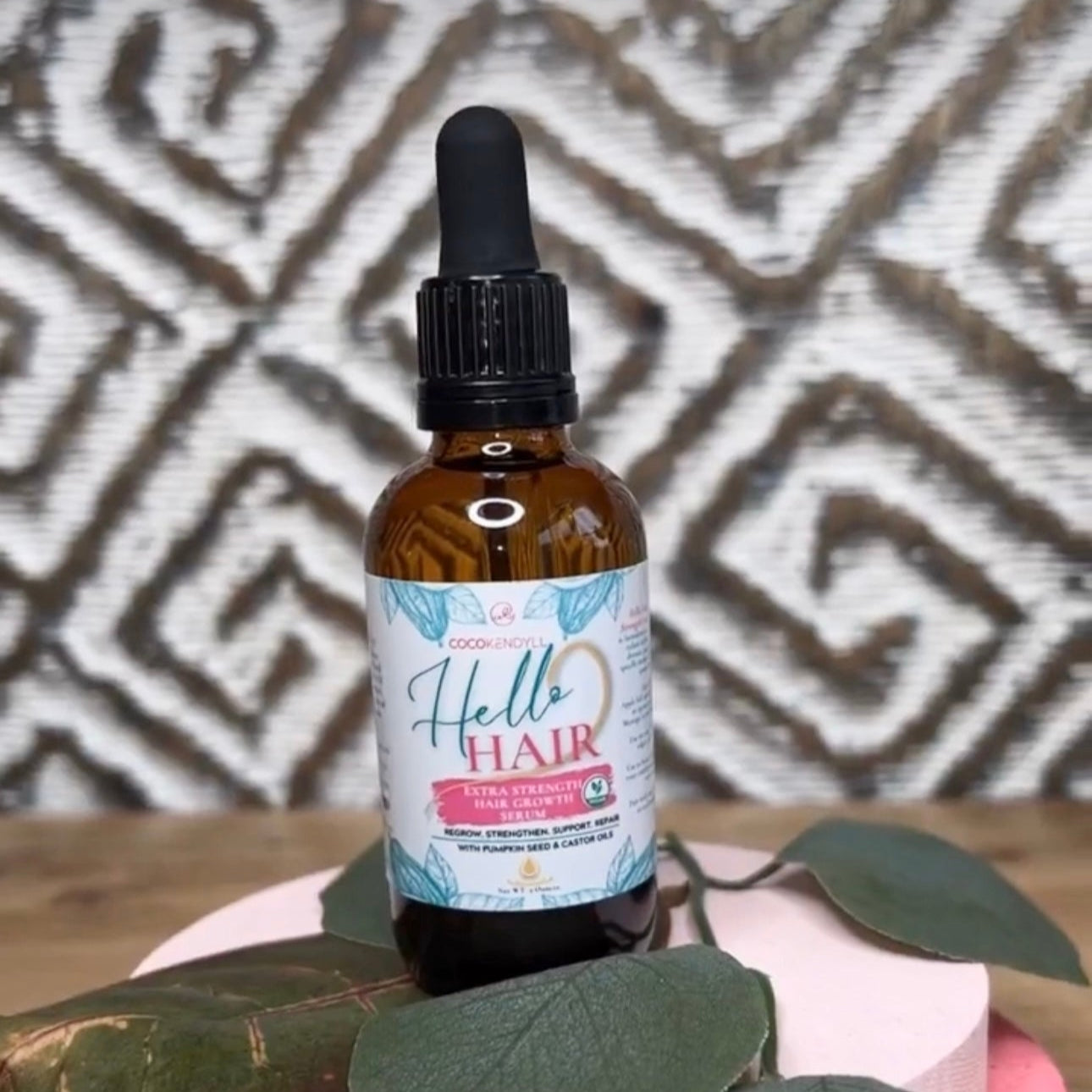 Coco Kendyll Hello Hair Extra Strength Organic Hair Regrowth Serum with dropper, 2 oz brown bottle, product photo on patterned background with green leaves.
